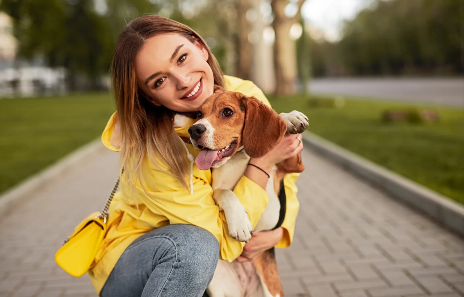 A smiling woman in a yellow raincoat crouching on a park path with her arms around a beagle