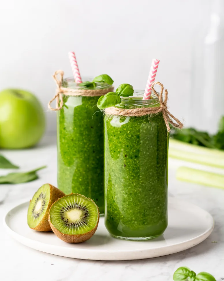 Two tall jars of green smoothie topped with basil and paper straws, beside a halved kiwi fruit, a green apple and celery