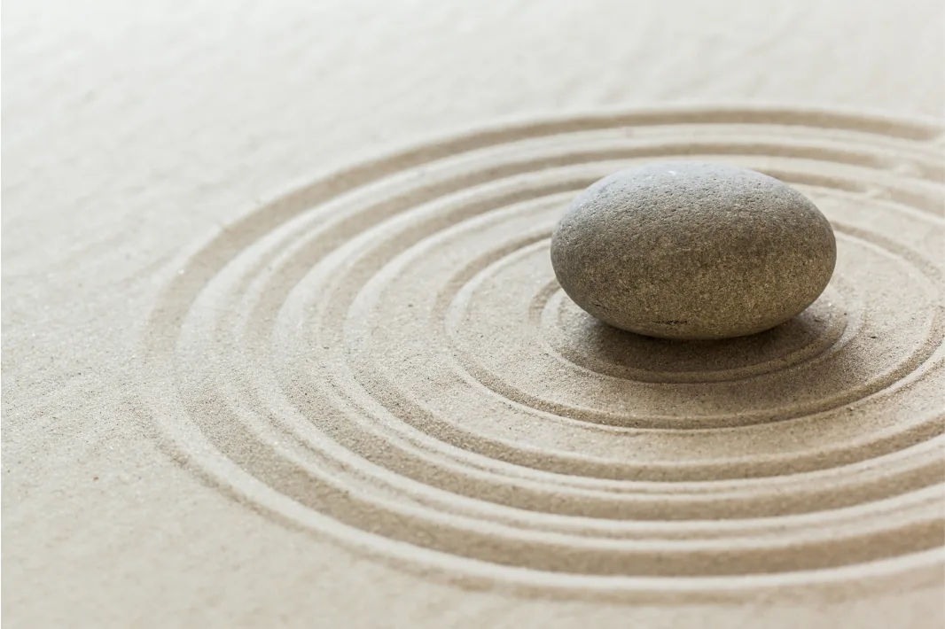 A single smooth grey stone resting at the centre of concentric circles raked into pale sand